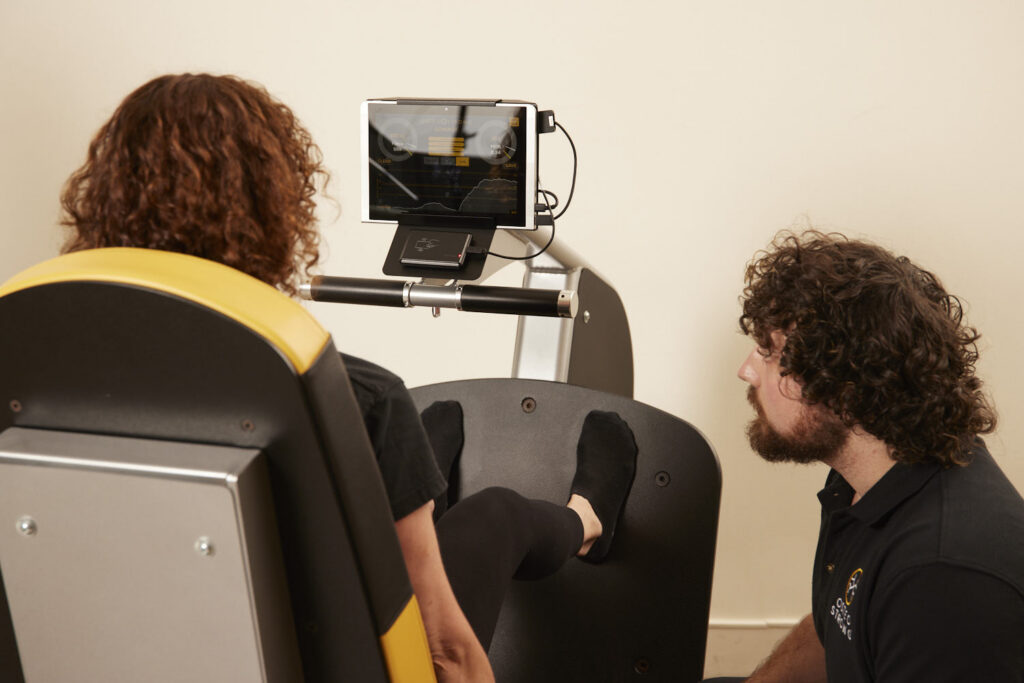 A woman uses an OsteoStrong machine while a trainer monitors her progress on a tablet screen